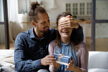 Smiling Millennial Hipster Man Covering Eyes Of Joyful Curious Young Wife, Giving Wrapped Box With Gift, Preparing Surprise Congratulating With Happy Birthday, Marriage Anniversary, Special Occasion.