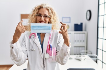 Poster - Middle age blonde woman wearing doctor uniform and medical mask winking looking at the camera with sexy expression, cheerful and happy face.