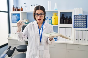 Poster - Young hispanic woman working at scientist laboratory with blood samples in shock face, looking skeptical and sarcastic, surprised with open mouth