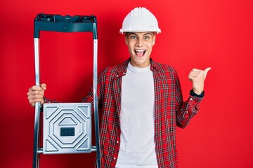 Canvas Print - Young hispanic man holding construction stairs wearing hardhat pointing thumb up to the side smiling happy with open mouth