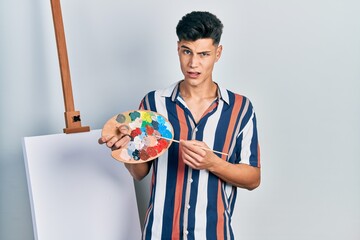 Canvas Print - Young hispanic man holding painter palette and paintbrush standing close to canvas skeptic and nervous, frowning upset because of problem. negative person.