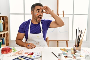 Poster - Young hispanic man with beard at art studio with painted face very happy and smiling looking far away with hand over head. searching concept.