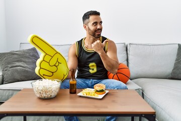 Poster - Young hispanic man with beard holding basketball ball cheering tv game pointing thumb up to the side smiling happy with open mouth