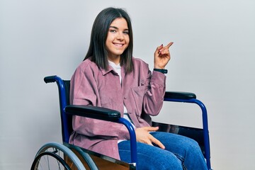 Poster - Young brunette woman sitting on wheelchair with a big smile on face, pointing with hand finger to the side looking at the camera.