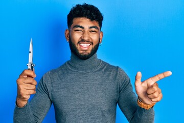 Wall Mural - Arab man with beard holding pocket knife smiling happy pointing with hand and finger to the side