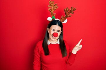 Poster - Young hispanic woman wearing deer christmas hat and red nose with a big smile on face, pointing with hand finger to the side looking at the camera.