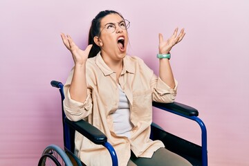 Poster - Young hispanic woman sitting on wheelchair crazy and mad shouting and yelling with aggressive expression and arms raised. frustration concept.