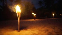 Row Of Natural Maiden Fire Torches On The Lonely Sandy Ocean Beach Glowing As A Part Of Ancient Ritual Dances. Dark Evening Handheld 4K Footage. Exotic Places And Traditions Concept Video.