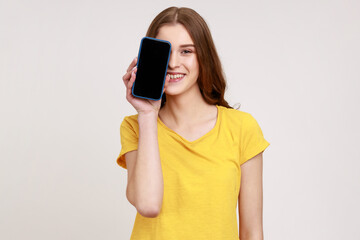 Wall Mural - Portrait of joyful teen girl with brown wavy hair in casual clothes smiling and covering half face with cell phone, looking playful and carefree. Indoor studio shot isolated on gray background.