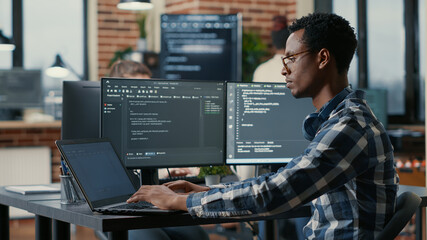 Wall Mural - Portrait of african american developer using laptop to write code sitting at desk with multiple screens parsing algorithm in software agency. Coder working on user interface using portable computer.