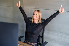 Happy Businesswoman Showing Thumps Up Sitting On Chair In Front Of Gray Wall At Office