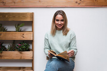 Smiling Woman With Book By Plants In Front Of Wall