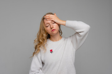 young blonde woman wears white sweater, touching her head and grimacing from pain, having severe headache, standing with migraine against gray background