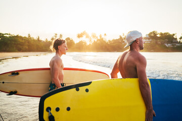 Wall Mural - Young teen boy with a surfboard with his father go to the sea for surfing. They are smiling and enjoying a beautiful sunset light. Family active vacation concept.