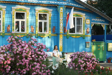 Wall Mural - Russian fashion woman, tourist girl in summer rose garden in Suzdal town, Russia. Look with collar with embroidery, skirt, hat, gloves. Street style, lady like style. Suzdal landmark, wooden house