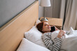 © dikushin - High-angle view of smiling young woman lying on cosy bed at home and talking mobile phone after waking up in morning at home. Cheerful redhead female having pleasant conversation on smartphone.