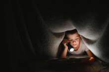 Boy With Flashlight Reading Book Under Blanket At Home