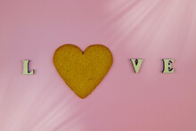 Inscription Love On A Pink Background. Top View Of Wooden Letters And Heart Shaped Gingerbread, Original Congratulations On Valentine's Day