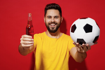 Young bearded man football fan in yellow t-shirt cheer up support favorite team hold give soccer ball bottle beer ale to you isolated plain dark red background studio portrait. Sport leisure concept.