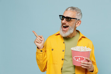 Wall Mural - Young elderly man in 3d glasses watch movie film hold bucket of popcorn point finger aside on workspace isolated on plain blue background studio portrait. People emotions in cinema lifestyle concept.