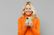 © ViDi Studio - Elderly smiling satisfied fun happy blonde caucasian woman 50s in orange turtleneck hold in hand use mobile cell phone isolated on plain grey color background studio portrait People lifestyle concept