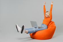 Full Body Elderly Smiling Happy Blonde Woman 50s In Orange Turtleneck Sit In Bag Chair Hold Use Work On Laptop Pc Computer Stretch Hands Finish Job Isolated On Plain Grey Background Studio Portrait