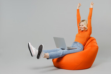 Full body elderly smiling happy blonde woman 50s in orange turtleneck sit in bag chair hold use work on laptop pc computer stretch hands finish job isolated on plain grey background studio portrait