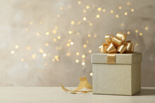 Beautiful Golden Gift Box On Light Table Against Blurred Festive Lights, Space For Text