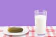 Pickled salty cucumber on a white saucer with a glass of milk on a red and white tablecloth on a purple background with copy space. Incompatible edible products. Concept of dyspepsia or upset stomach