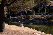 Two People Sit On Park Bench Watching Activities Across A Stream