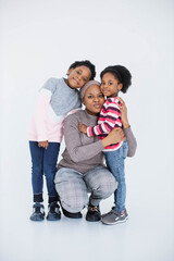 Wall Mural - Portrait of happy african islamic woman embracing her little daughters while posing in studio with white background. Two cute sisters spending time with their caring lovely mother.