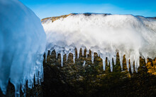 The White Plate Of Ice And Icicles Over The Dark Boulders On The Blue Sky Background