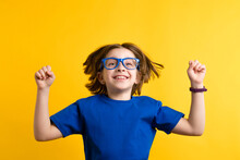 Little Girl Winner 8-10 Years Old In Basic Blue T-shirt And Glasses On Yellow Background. Gender Neutral Child Having Fun And Clenched Fists In Victorious Jump. Childhood Emotions, Facial Expression.