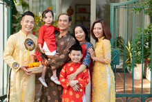 Smiling Grandparents, Parents And Children In Traditional Ao Dai Dresses Standing In Front Of House Ready For Tet Celebration