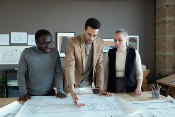 Poster - Young biracial businessman making presentation of sketch on unrolled blueprint to his colleagues at meeting