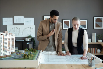Poster - Two elegant architects or engineers discussing sketch on blueprint while standing by large table with house layout