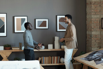 Poster - Two young intercultiral businessmen discussing working points while standing in front of each other against wall with paintings
