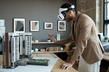 Poster - Young elegant architect in vr headset standing by table with house layout during presentation of his new project