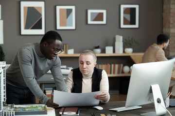 Poster - Two intercultural man and woman discussing working document by workplace against their colleague in office environment