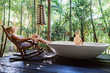 Happy baby boy have fun in bathtub, young mother relax on rocking chair in outside bathroom on veranda with beautiful tropical garden view at luxury villa. Family summer vacation with kids.