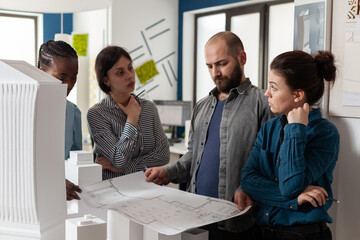 Poster - Team of engineers looking at architect holding blueprints for construction project in modern architectural office. Professional engineers analyzing paper plans for building in front of foam maquette.