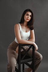 Carefree young woman in smart casual wear sitting on a chair, posing in studio over gray background. Relaxed and beautiful young woman