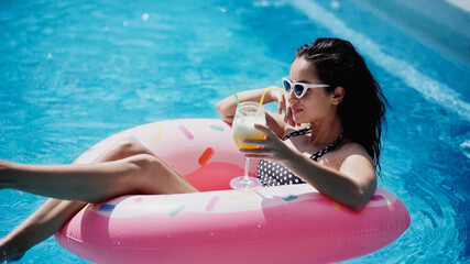 Wall Mural - cheerful woman in sunglasses holding glass with cocktail and swimming on inflatable ring in pool