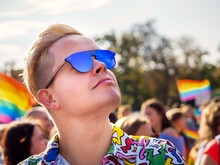 Portrait Of A Gay Person On LGBT Pride Parade With Waving Rainbow Flags On Background In European City In A Sunny Day