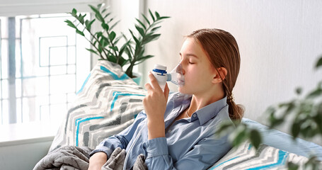 Teenage girl makes inhalation with a nebulizer equipment. Sick child holding inhalator in hand and breathes through an inhaler at home