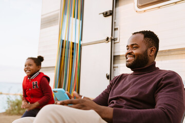 Wall Mural - Black man using cellphone while resting with his daughter by trailer