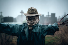 Terrible Scarecrow In Dark Cloak And Dirty Hat Stands Alone On A Cemetery Background. Halloween Concept
