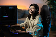 Portrait Of A Laughing Young Girl Gamer With Headphones Sitting At The Computer On A Comfortable Chair, Hands On The Backlit Keyboard And Mouse, Dark Room Illuminated By Led Blue Orange Lighting