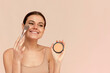 A beautiful woman applies powder using a cosmetic brush on her face skin on a pink background.