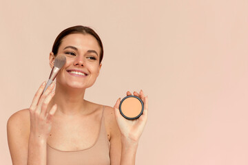 Wall Mural - A beautiful woman applies powder using a cosmetic brush on her face skin on a pink background.